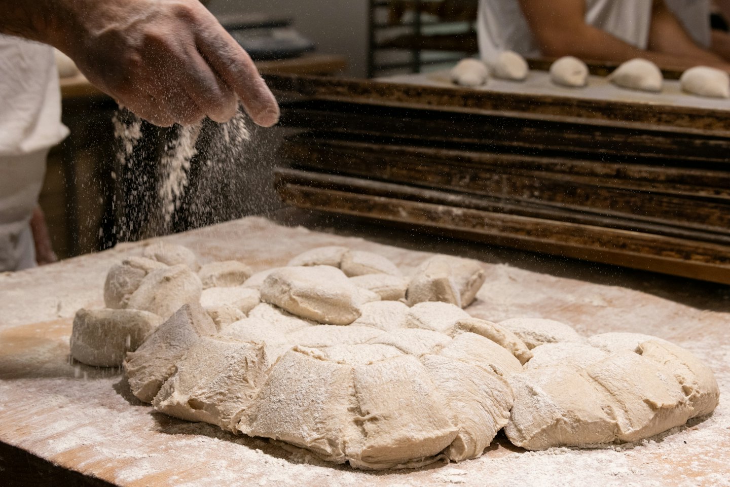 A baker sprinkles flour onto dough on a wooden table.
